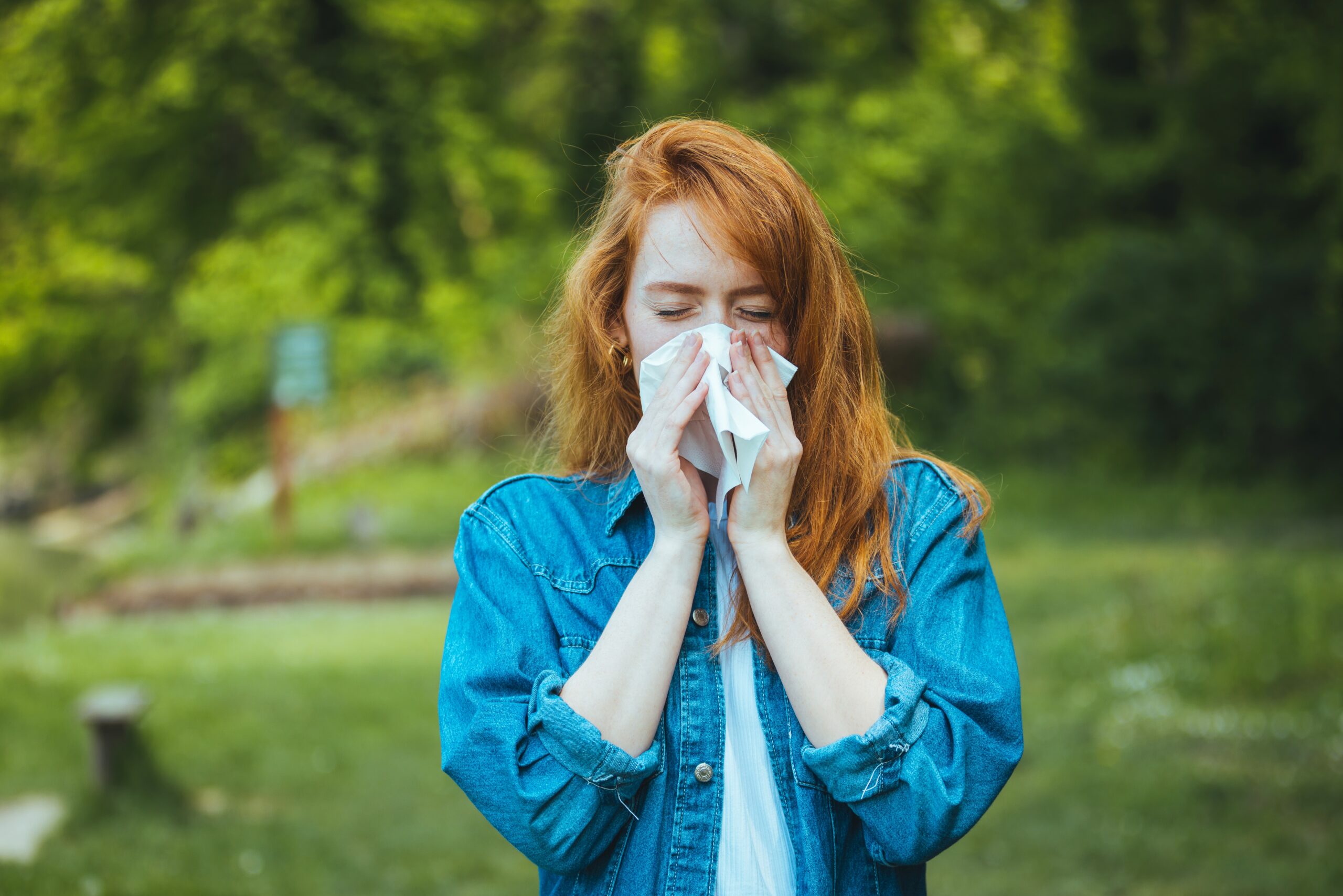 A young lady dressed for mild weather blows her nose, surrounded by greenery on a sunny day